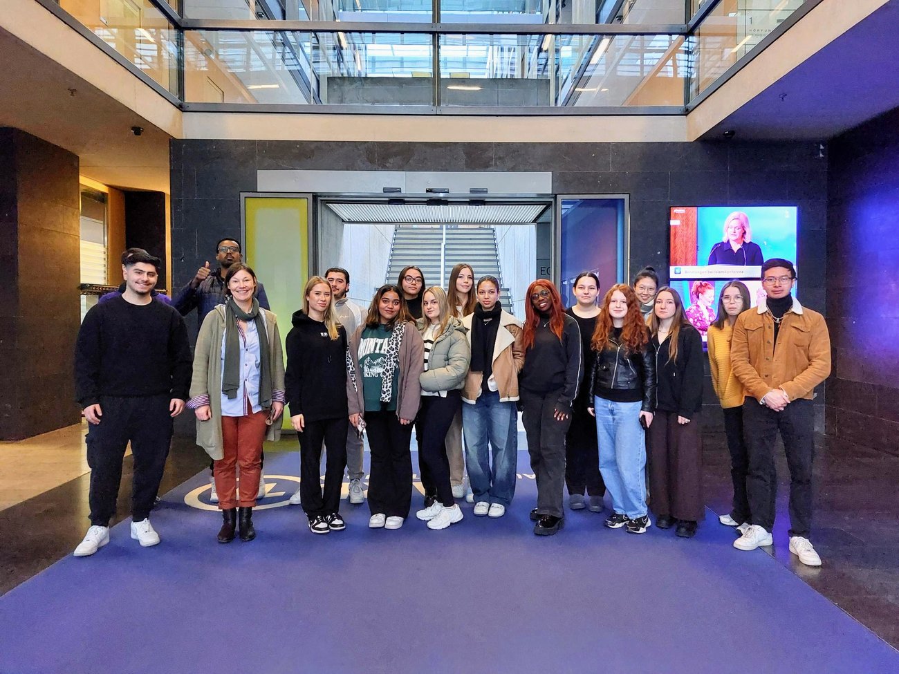 Gruppe von Schülerinnen und Schülern auf einem blauen Teppich mit einem Bildschirm im rechten Hintergrund