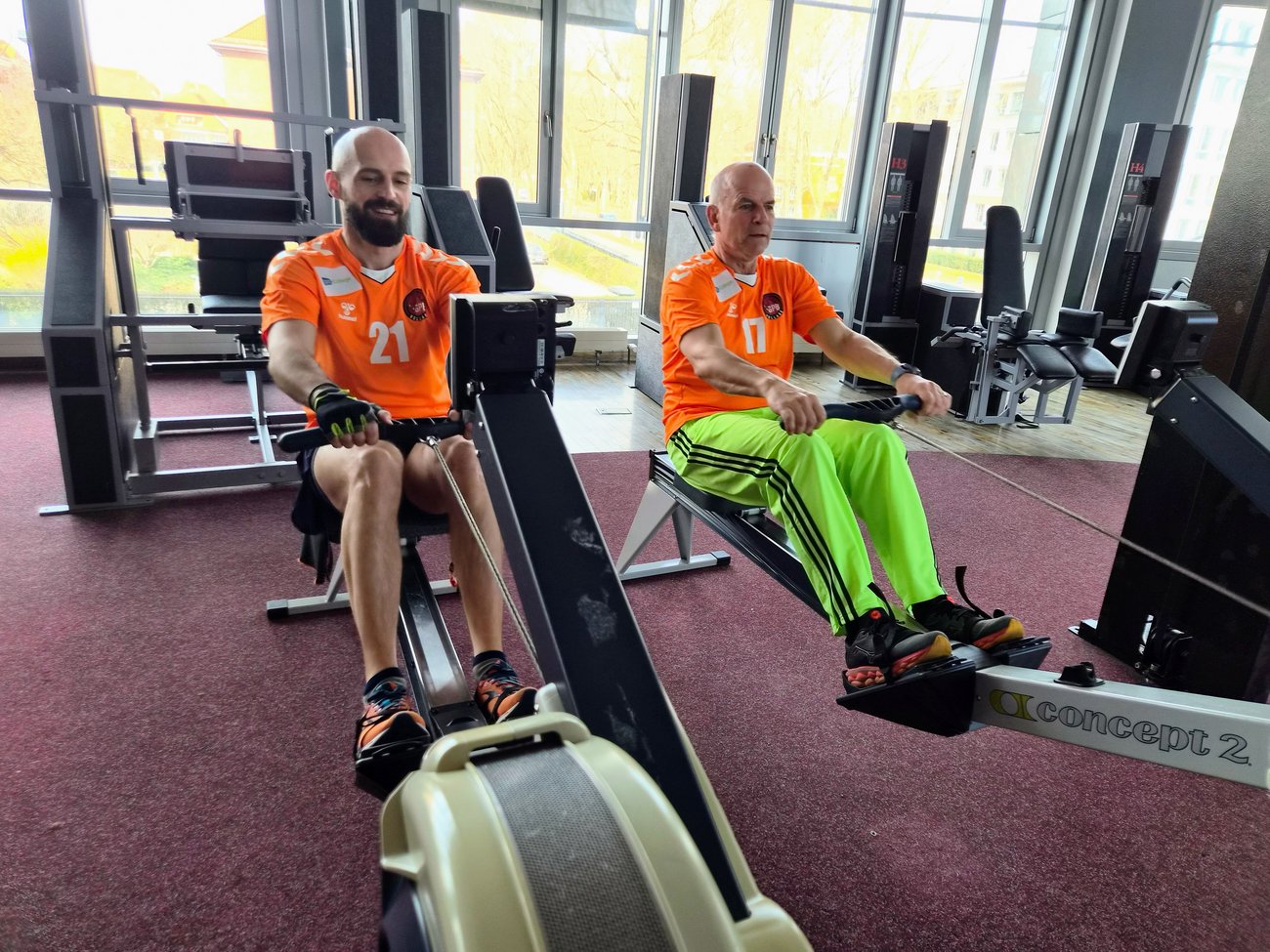 zwei Männer in orangen Trikots an der Rudermaschine