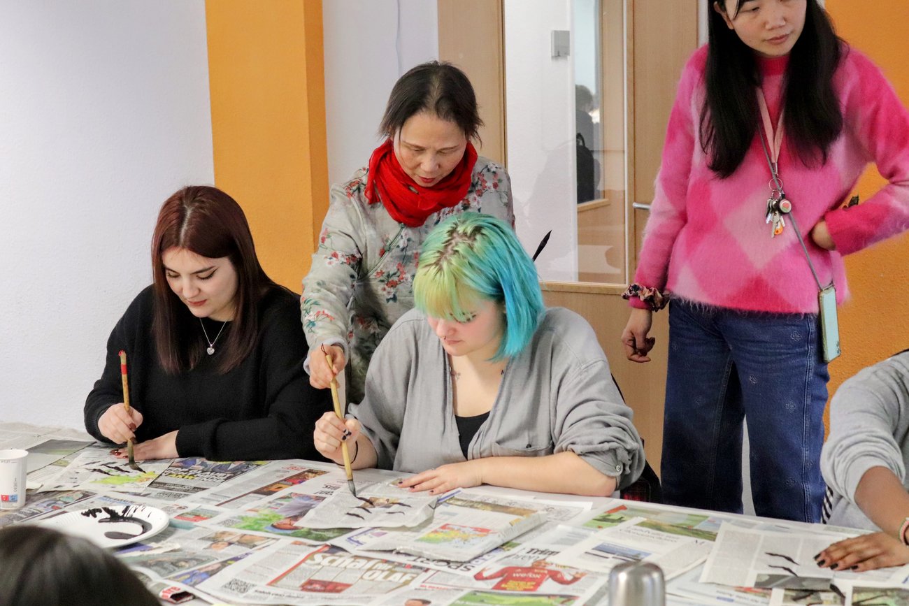 eine Lehrerin zeigt zwei Schülerinnen die richtige Pinselhaltung beim Malen der asiatischen Schriftzeichen