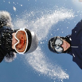 zwei Köpfe mit Skibrille und Helm und in die Höhe geworfener Schnee