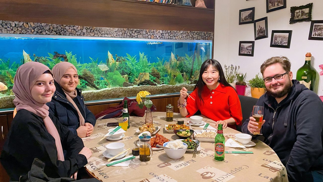 eine Lehrerin mit drei Schülern und asiatischem Essen auf dem Tisch, im Hintergrund ein großes Aquarium