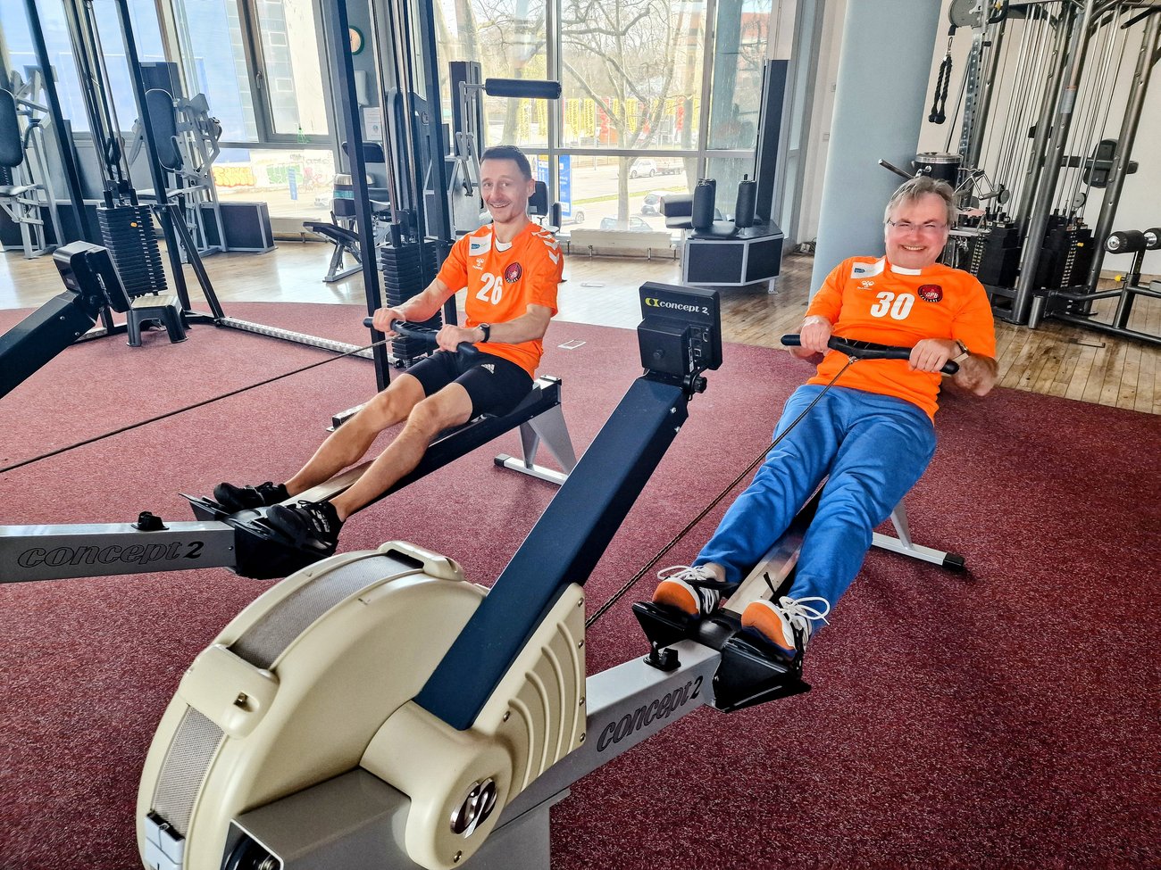 zwei Männer in orangen Trikots auf der Rudermaschine