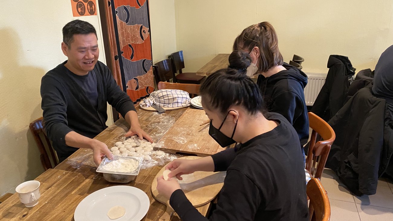 zwei Schülerinnen formen Teigstücke, ein asiatischer Koch reicht den Teig